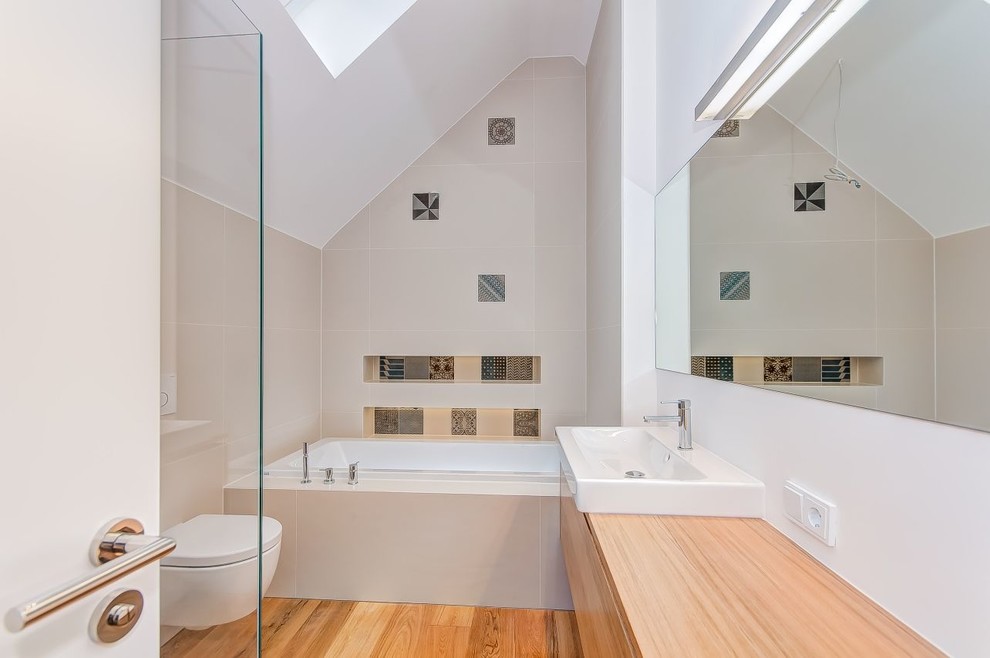 Inredning av ett modernt litet badrum, med släta luckor, skåp i mellenmörkt trä, ett badkar i en alkov, en vägghängd toalettstol, flerfärgad kakel, beige väggar, mellanmörkt trägolv, ett fristående handfat och träbänkskiva
