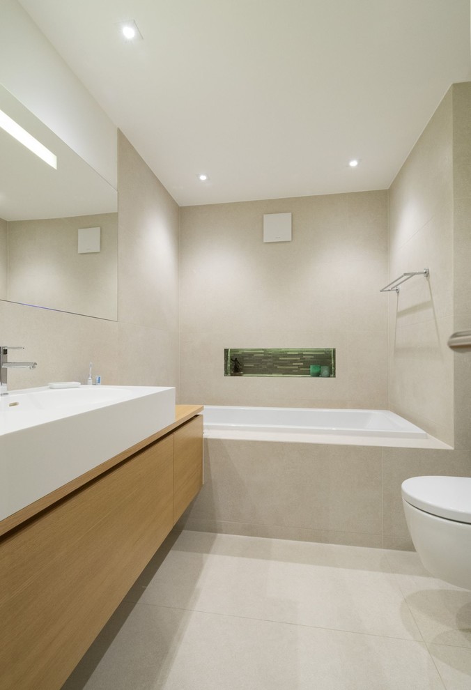Modern inredning av ett litet badrum, med släta luckor, skåp i mellenmörkt trä, ett badkar i en alkov, beige kakel, ett fristående handfat, träbänkskiva, stenkakel, beige väggar och kalkstensgolv