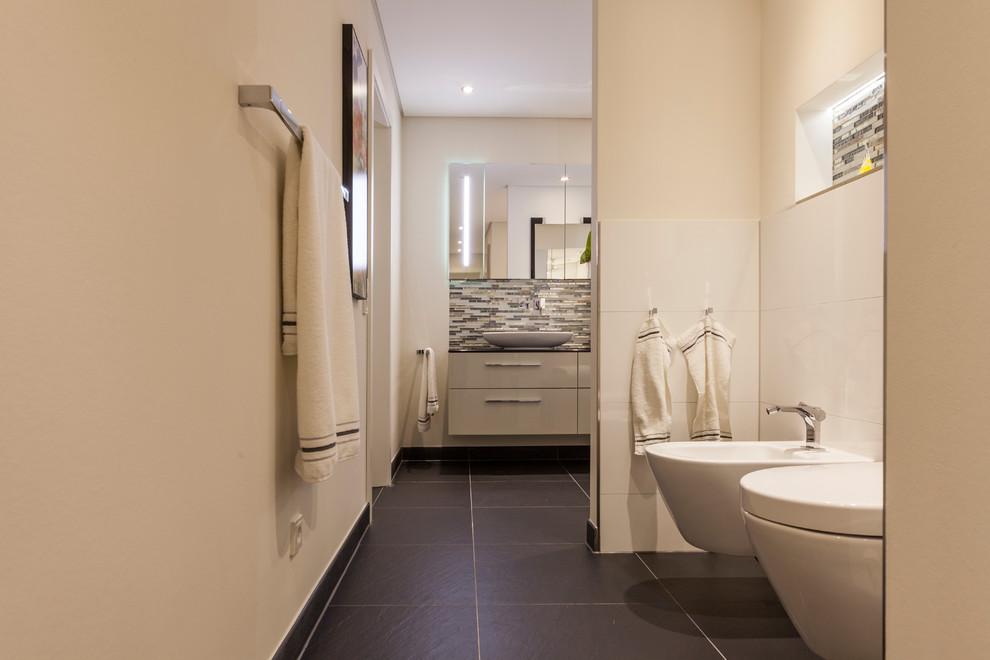 This is an example of a large contemporary bathroom in Cologne with flat-panel cabinets, beige cabinets, beige tiles, brown tiles, grey tiles, beige walls, a vessel sink, solid surface worktops, a bidet, matchstick tiles, cement flooring and brown floors.