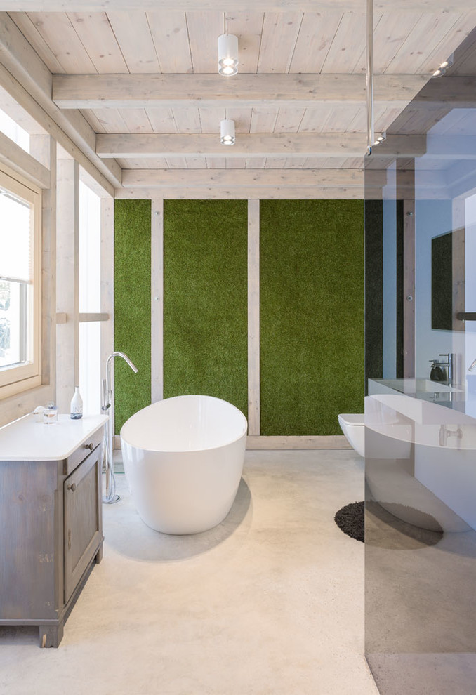 Inredning av ett modernt stort en-suite badrum, med en kantlös dusch, en vägghängd toalettstol, gröna väggar, betonggolv, ett väggmonterat handfat, med dusch som är öppen, skåp i ljust trä, ett fristående badkar, grått golv och luckor med infälld panel