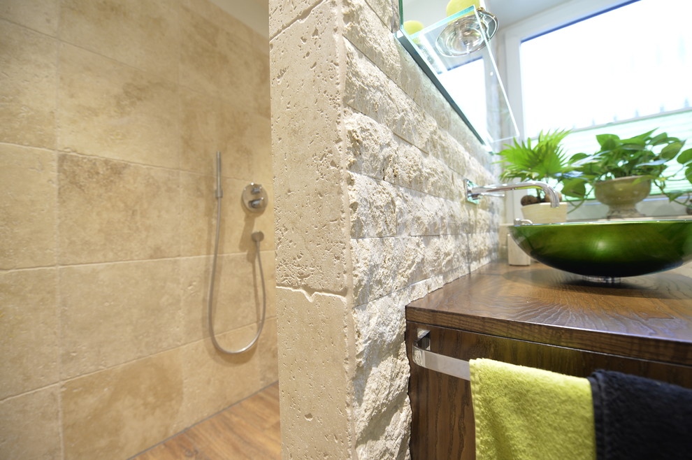 Exemple d'une salle d'eau tendance en bois foncé de taille moyenne avec une douche à l'italienne, WC suspendus, un carrelage beige, des dalles de pierre, un mur beige, une vasque, un plan de toilette en verre, un sol marron et aucune cabine.