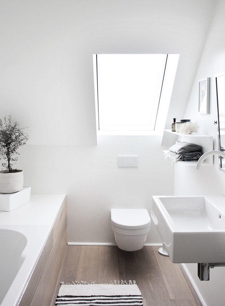 Modelo de cuarto de baño principal minimalista pequeño con paredes blancas, suelo de madera clara, lavabo suspendido, bañera encastrada, sanitario de dos piezas y suelo marrón