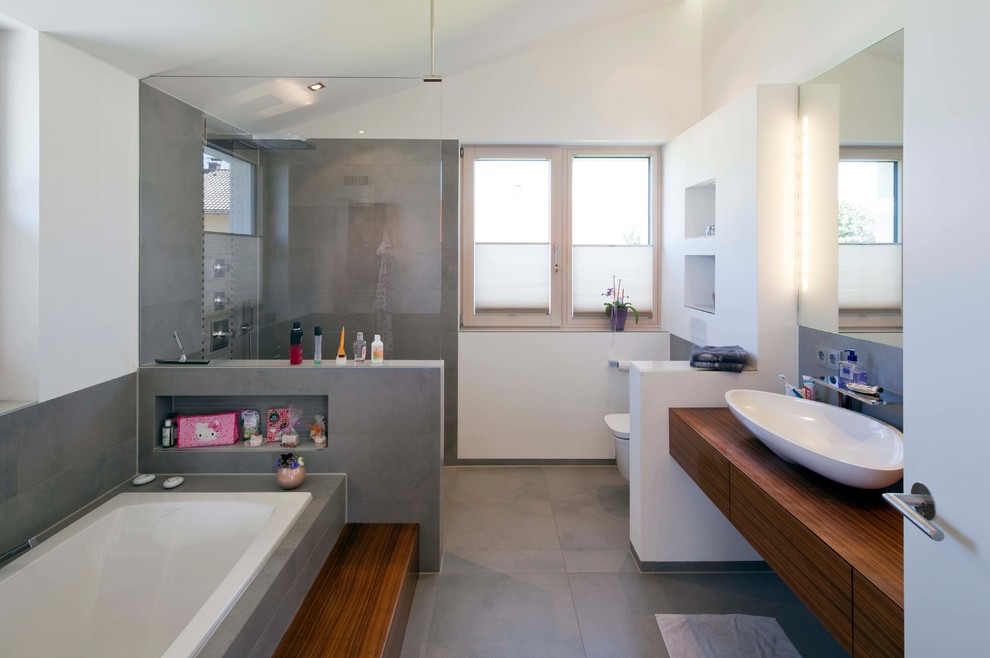 Modern inredning av ett stort brun brunt badrum med dusch, med släta luckor, skåp i mellenmörkt trä, ett platsbyggt badkar, en öppen dusch, en vägghängd toalettstol, grå kakel, cementkakel, vita väggar, cementgolv, ett fristående handfat, träbänkskiva, grått golv och med dusch som är öppen