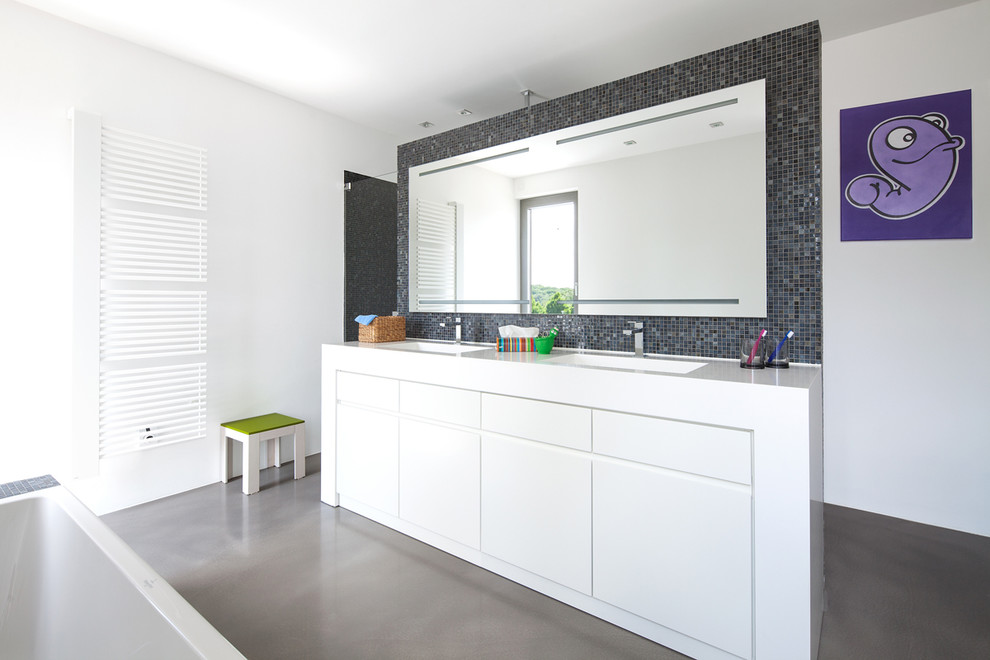 Diseño de cuarto de baño contemporáneo grande con armarios con paneles lisos, puertas de armario blancas, baldosas y/o azulejos negros, baldosas y/o azulejos en mosaico, paredes blancas, bañera encastrada, lavabo integrado y espejo con luz