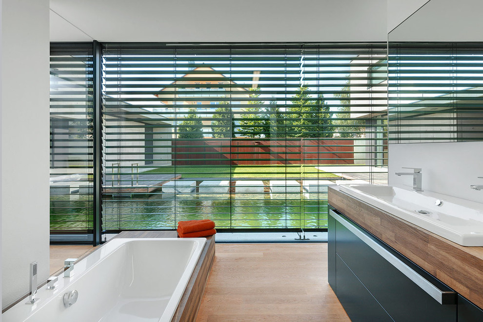 ニュルンベルクにあるコンテンポラリースタイルのおしゃれな浴室 (フラットパネル扉のキャビネット、中間色木目調キャビネット、ドロップイン型浴槽、白い壁、無垢フローリング、オーバーカウンターシンク、木製洗面台) の写真