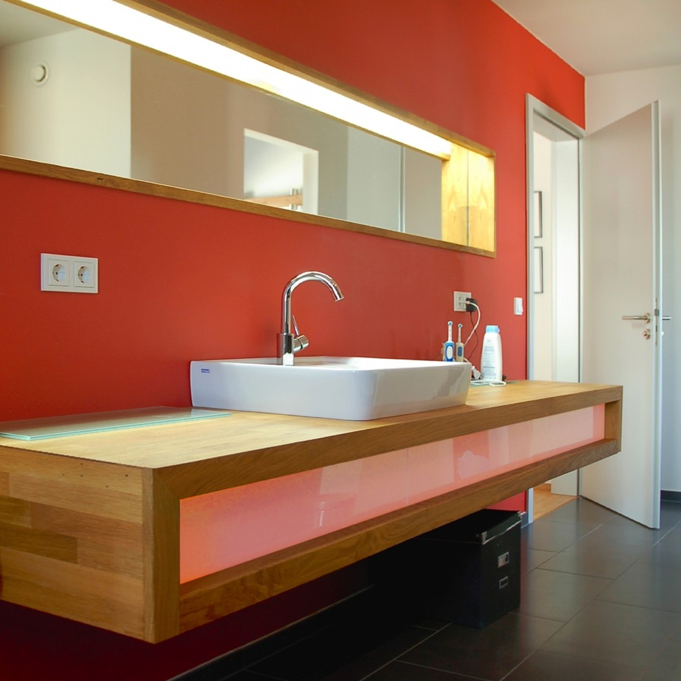 This is an example of a medium sized contemporary shower room bathroom in Nuremberg with glass-front cabinets, medium wood cabinets, a freestanding bath, a built-in shower, a wall mounted toilet, red tiles, red walls, slate flooring, a vessel sink, solid surface worktops, black floors, an open shower and beige worktops.