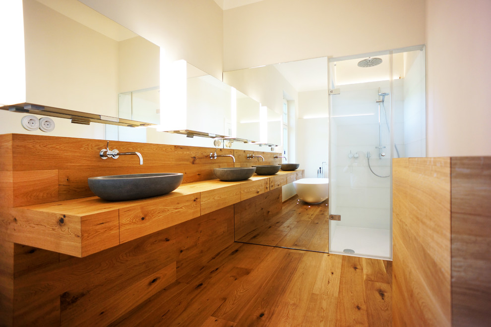 Modern inredning av ett stort badrum, med ett fristående handfat, skåp i mellenmörkt trä, träbänkskiva, ett fristående badkar, mellanmörkt trägolv, släta luckor, en dusch i en alkov, vit kakel, keramikplattor och beige väggar