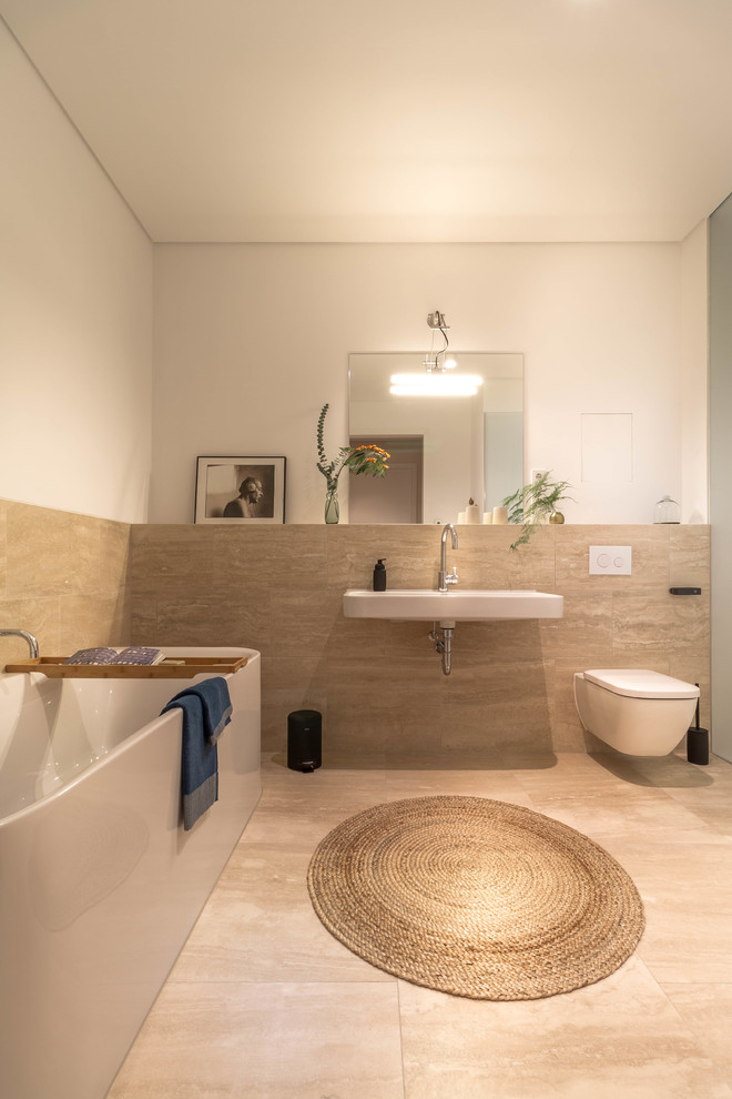 Cette photo montre une salle d'eau tendance de taille moyenne avec une baignoire indépendante, WC suspendus, un carrelage beige, des dalles de pierre, un sol en calcaire, un lavabo suspendu, un sol beige et un mur blanc.