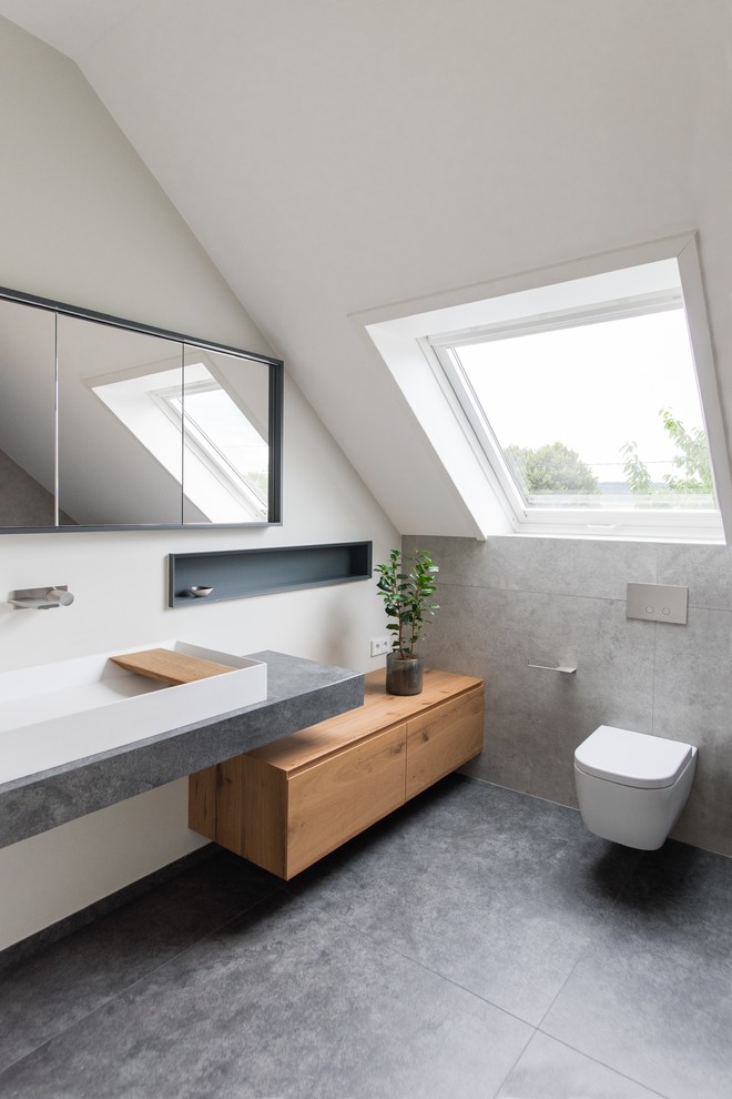 Bild på ett mellanstort funkis badrum med dusch, med skåp i ljust trä, en kantlös dusch, vita väggar, släta luckor, en vägghängd toalettstol, beige kakel, cementkakel, cementgolv, grått golv och ett avlångt handfat