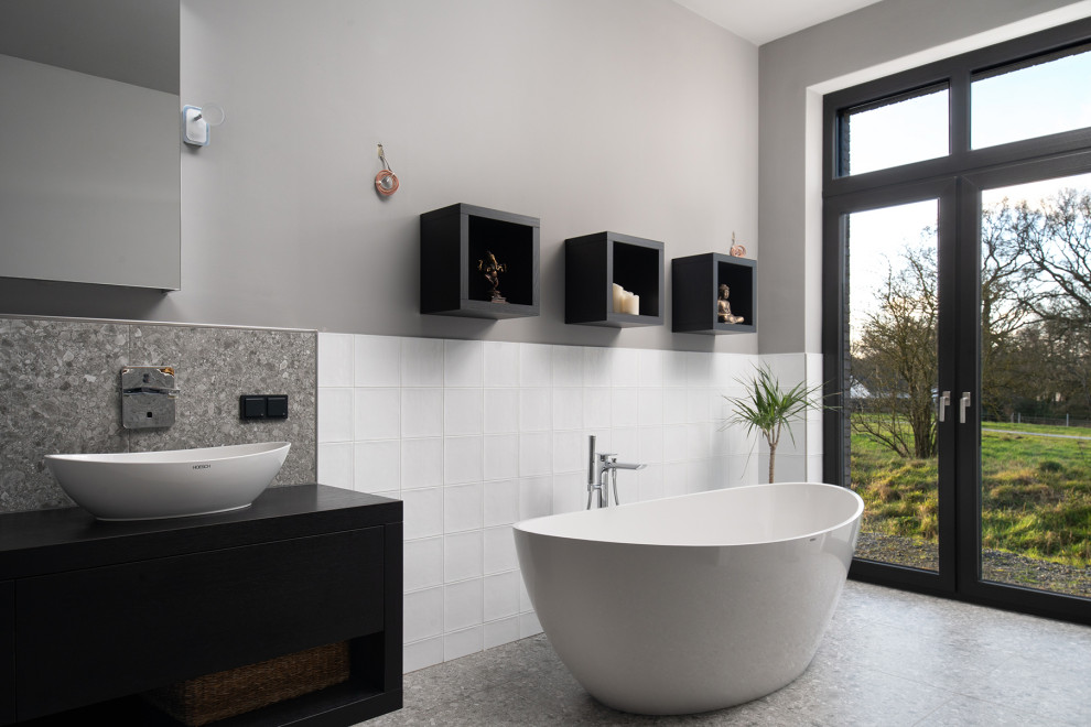 Bild på ett stort funkis svart svart en-suite badrum, med släta luckor, svarta skåp, ett fristående badkar, vit kakel, porslinskakel, grå väggar, terrazzogolv, ett fristående handfat och grått golv