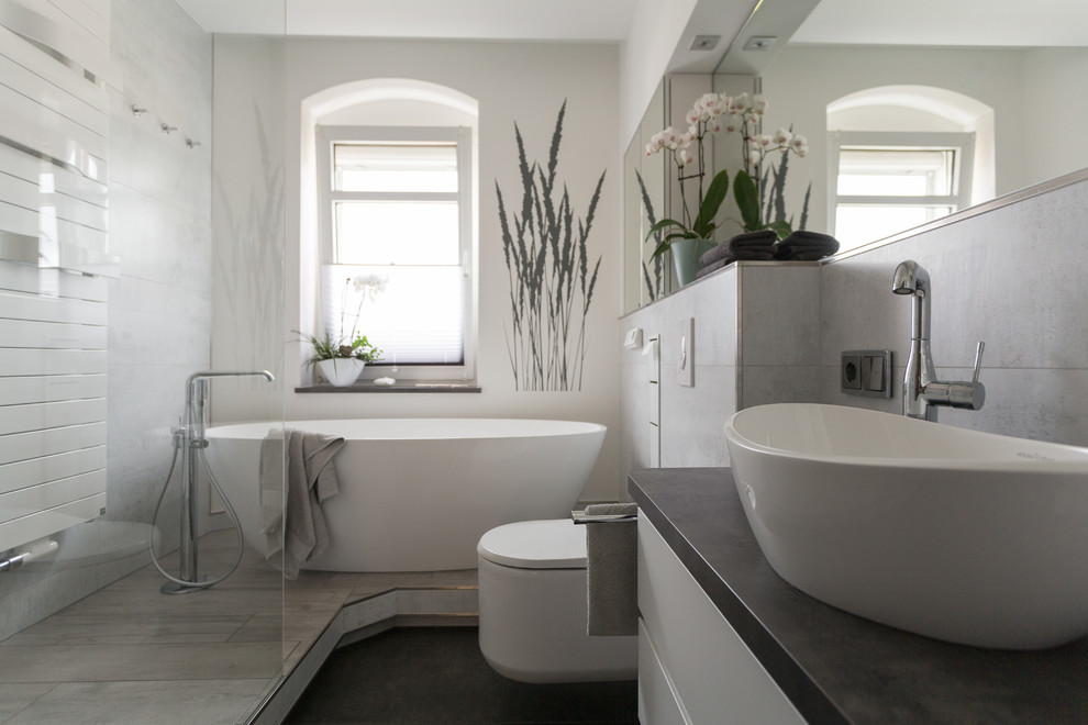 Foto på ett mellanstort funkis svart badrum med dusch, med släta luckor, vita skåp, ett fristående badkar, en kantlös dusch, en vägghängd toalettstol, grå kakel, vita väggar, ett fristående handfat, beiget golv och dusch med gångjärnsdörr