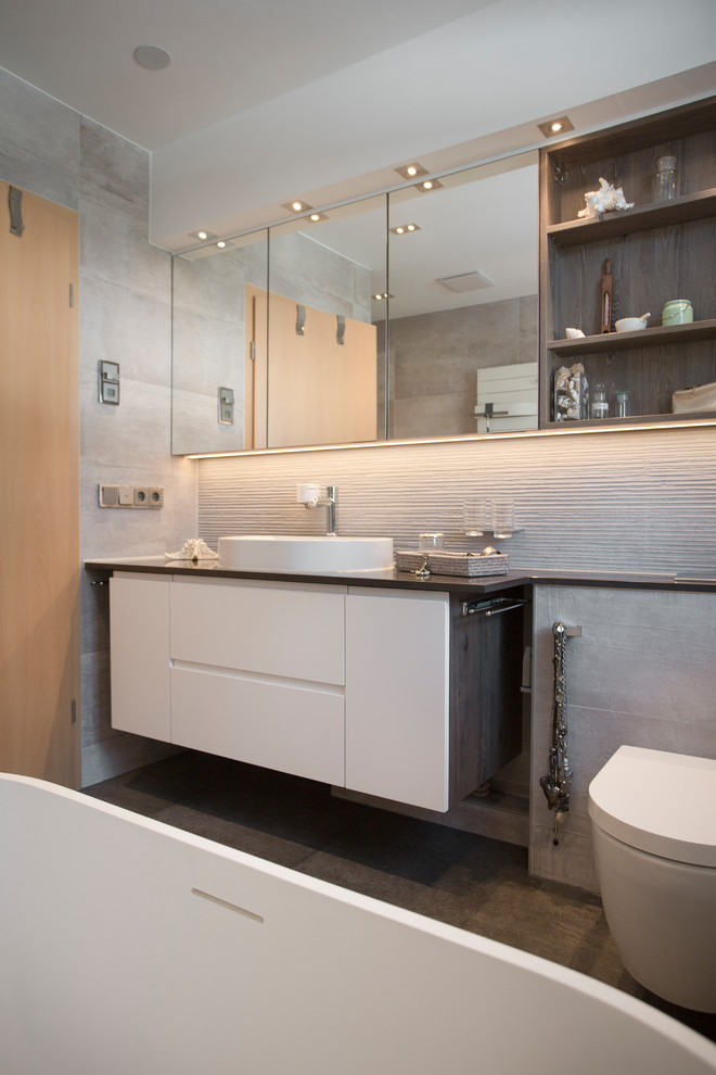Inredning av ett modernt mellanstort brun brunt en-suite badrum, med släta luckor, vita skåp, ett fristående badkar, en vägghängd toalettstol, beige kakel, cementkakel, vita väggar, cementgolv, ett fristående handfat och grått golv