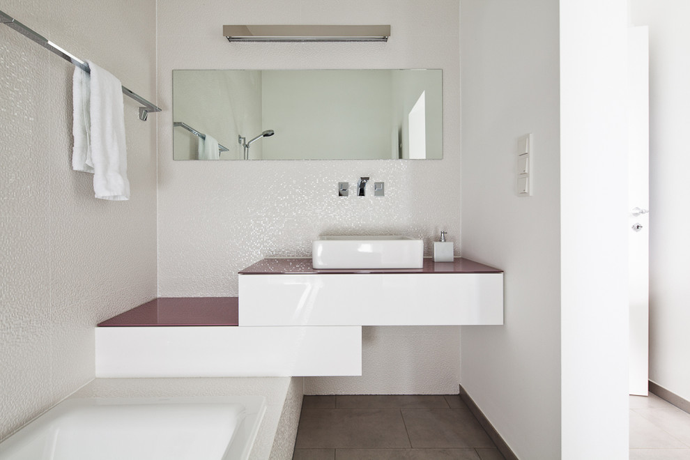 フランクフルトにある小さなコンテンポラリースタイルのおしゃれな浴室 (ベッセル式洗面器、フラットパネル扉のキャビネット、白いキャビネット、白い壁、ドロップイン型浴槽、シャワー付き浴槽	、白いタイル、ガラスの洗面台) の写真