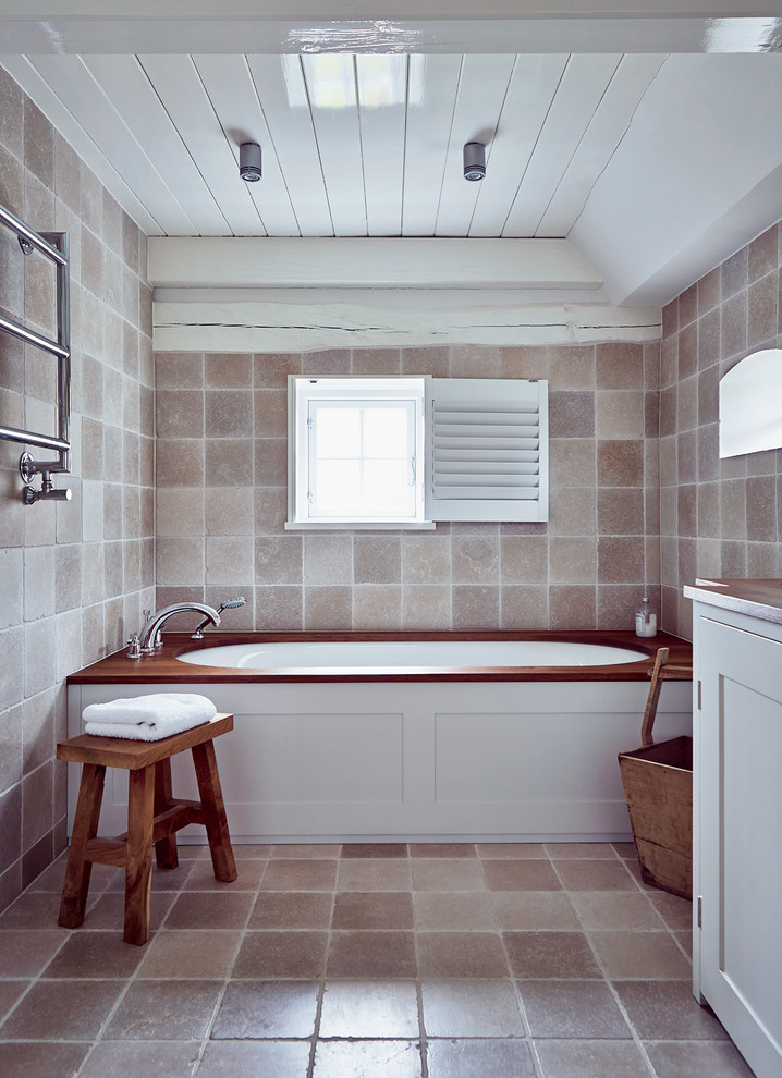 Cette image montre une petite salle de bain rustique avec une baignoire encastrée, un carrelage de pierre, un placard à porte shaker, des portes de placard blanches, un carrelage marron, un mur marron et un plan de toilette en bois.
