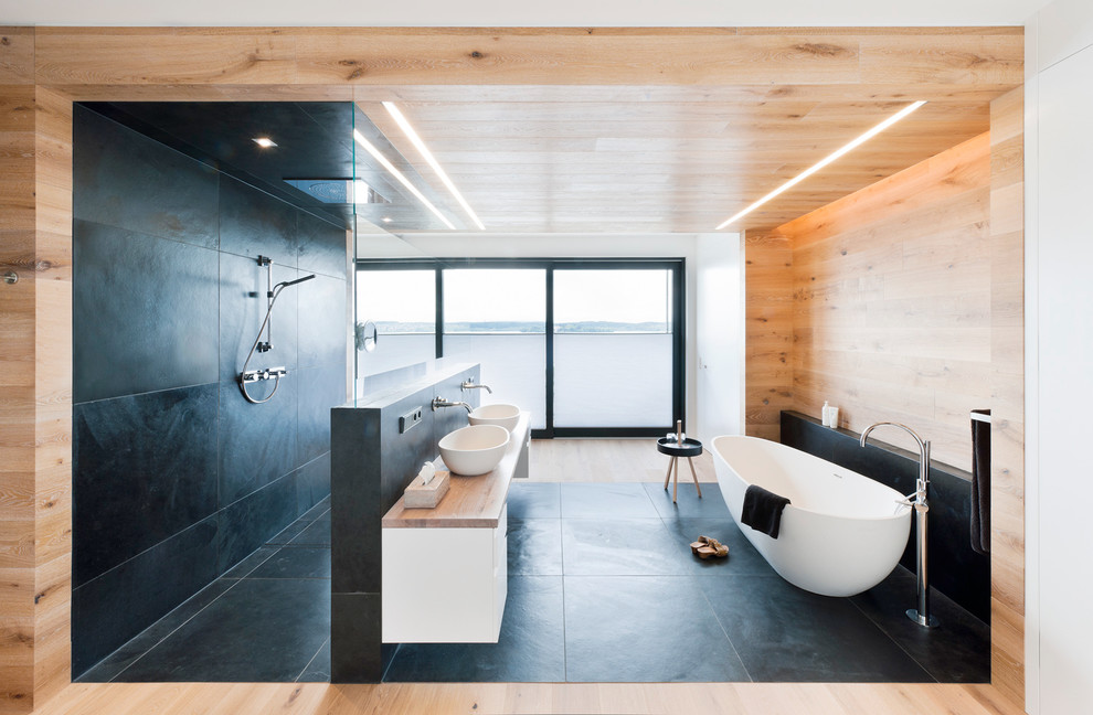 Modern inredning av ett stort badrum, med släta luckor, vita skåp, ett fristående badkar, en kantlös dusch, flerfärgade väggar, träbänkskiva, svart golv och med dusch som är öppen