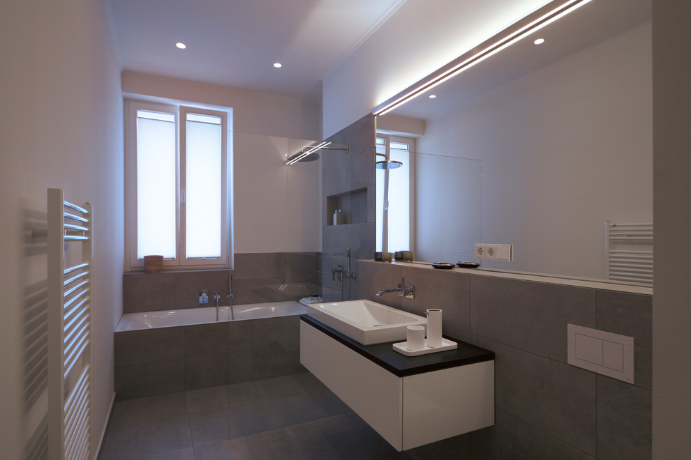 Modelo de cuarto de baño contemporáneo de tamaño medio con puertas de armario blancas, bañera encastrada, ducha abierta, sanitario de pared, baldosas y/o azulejos grises, paredes blancas, aseo y ducha, lavabo sobreencimera, suelo gris y ducha abierta