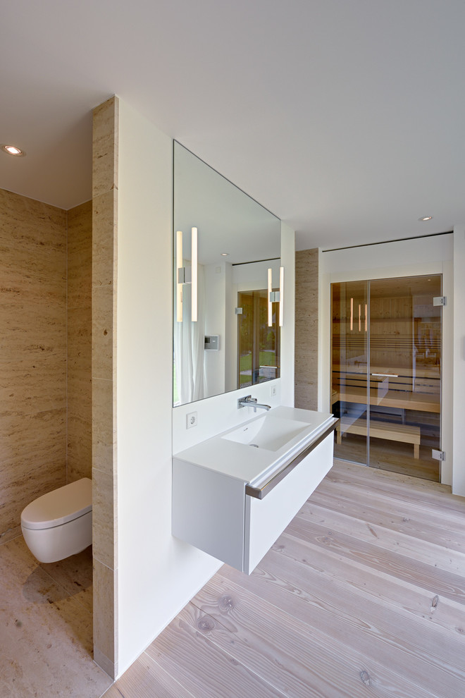 Inspiration for a large contemporary sauna bathroom in Berlin with flat-panel cabinets, white cabinets, a wall mounted toilet, beige tiles, stone slabs, white walls, light hardwood flooring and an integrated sink.