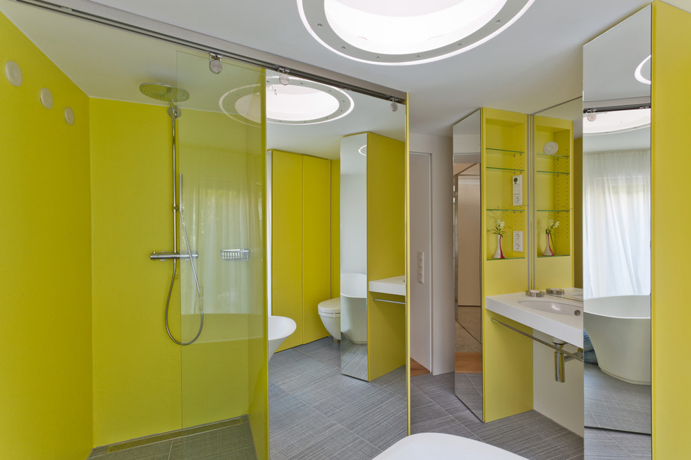 Modern inredning av ett mellanstort badrum, med ett väggmonterat handfat, grå kakel, gula väggar, klinkergolv i porslin, släta luckor, gula skåp, ett fristående badkar, en kantlös dusch och en vägghängd toalettstol