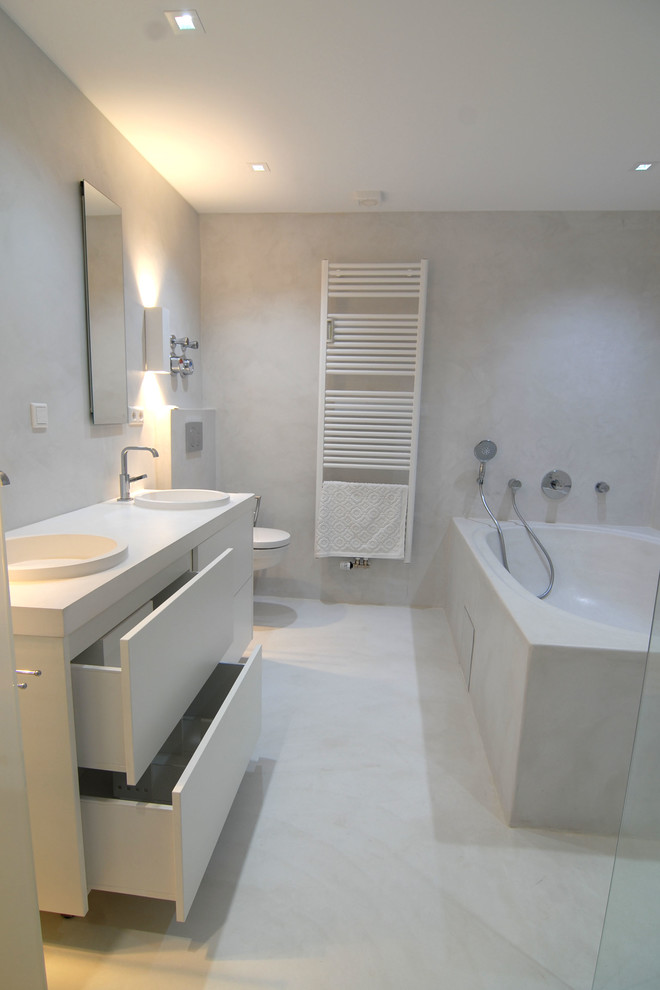 Foto på ett funkis badrum, med vita skåp, ett hörnbadkar, en kantlös dusch, en vägghängd toalettstol, vit kakel, vita väggar och ett avlångt handfat