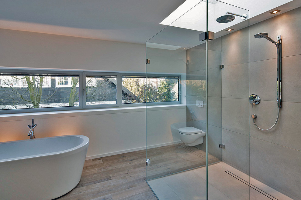 Großes Modernes Badezimmer mit freistehender Badewanne, bodengleicher Dusche, Wandtoilette, grauen Fliesen, Steinplatten, weißer Wandfarbe und braunem Holzboden in Düsseldorf