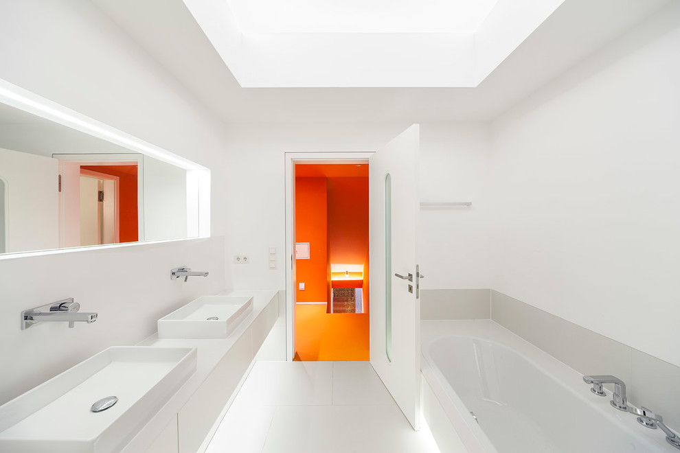 Cette image montre une salle d'eau minimaliste de taille moyenne avec un placard à porte plane, des portes de placard blanches, une baignoire posée, un mur blanc, une vasque, un sol blanc et un plan de toilette blanc.