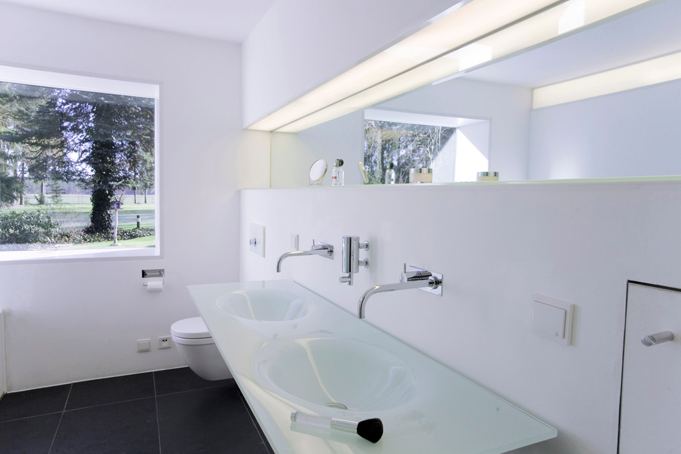 Imagen de cuarto de baño contemporáneo de tamaño medio con sanitario de pared, paredes blancas, lavabo integrado, encimera de vidrio y espejo con luz