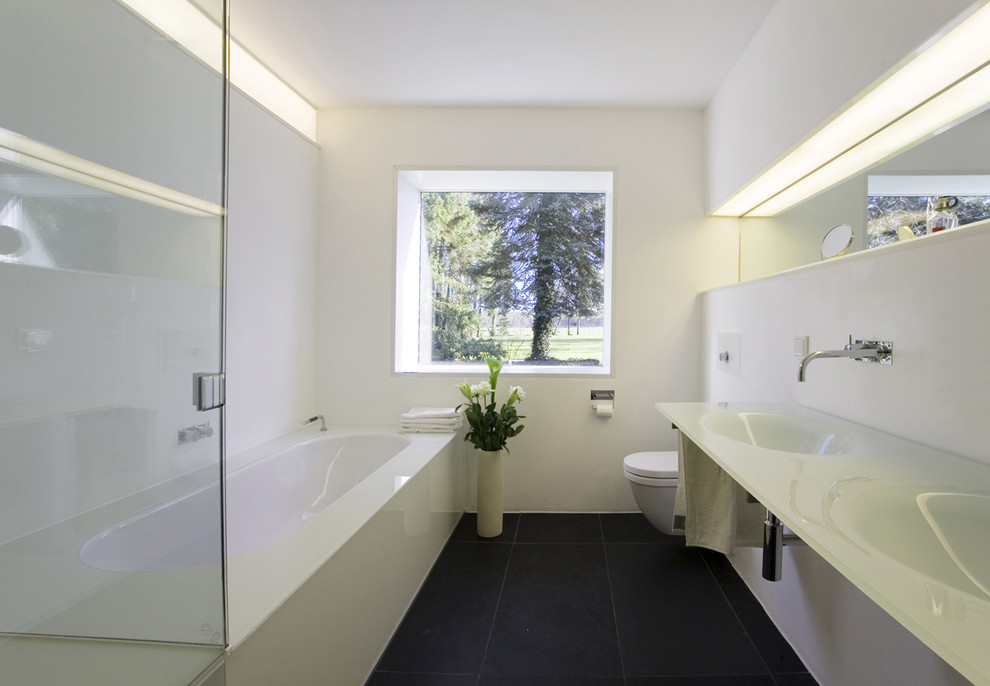 Свежая идея для дизайна: ванная комната среднего размера в современном стиле с монолитной раковиной, полновстраиваемой ванной, инсталляцией, белыми стенами, стеклянной столешницей и зеркалом с подсветкой - отличное фото интерьера