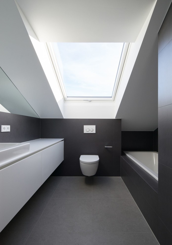 Kleines Modernes Badezimmer mit flächenbündigen Schrankfronten, weißen Schränken, Wandtoilette, grauen Fliesen, schwarzen Fliesen, weißer Wandfarbe, Aufsatzwaschbecken und Einbaubadewanne in München