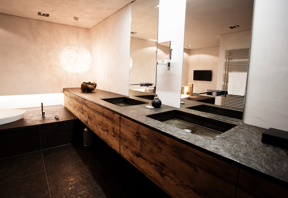 Diseño de cuarto de baño rural de tamaño medio con armarios con paneles lisos, puertas de armario de madera oscura, baldosas y/o azulejos negros, paredes blancas, suelo de pizarra, lavabo integrado y encimeras negras