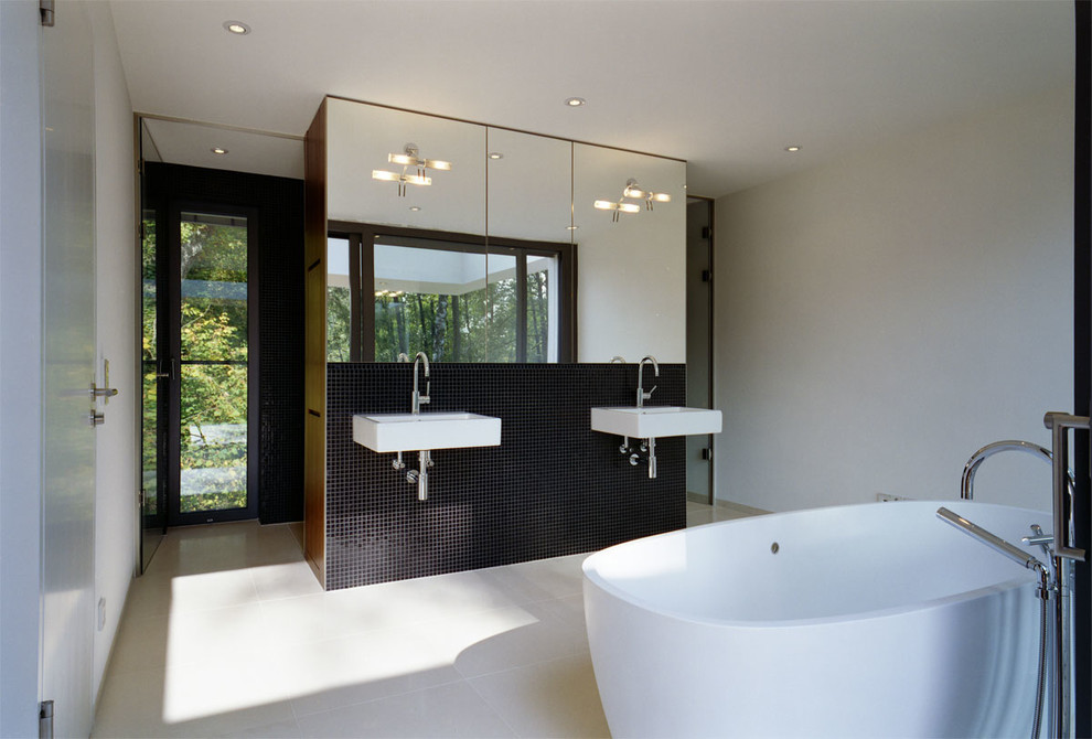 Large modern bathroom in Munich with a freestanding bath, black tiles, mosaic tiles, white walls and a wall-mounted sink.