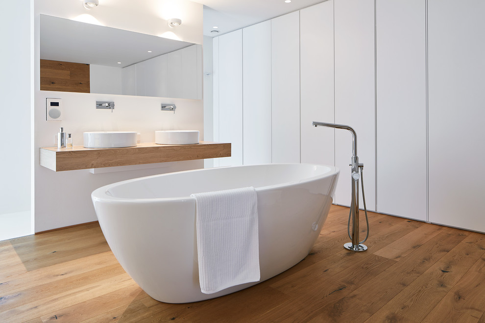 На фото: большая главная ванная комната в стиле модернизм с плоскими фасадами, белыми фасадами, отдельно стоящей ванной, столешницей из дерева, белыми стенами, светлым паркетным полом, коричневым полом, консольной раковиной и зеркалом с подсветкой