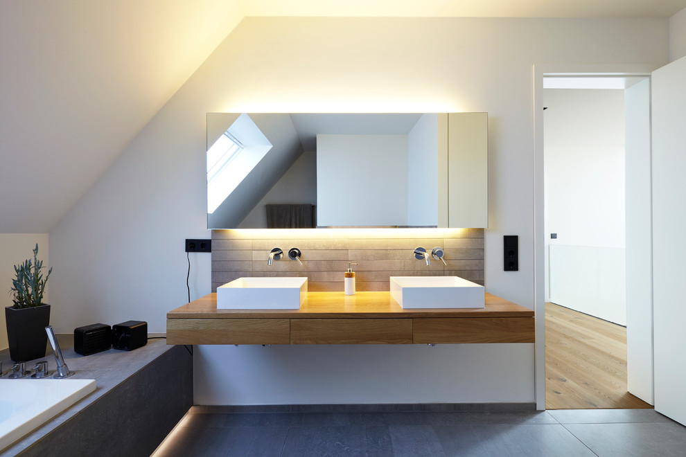 Diseño de cuarto de baño escandinavo, de tamaño medio, con lavabo sobreencimera, armarios con paneles lisos, puertas de armario de madera oscura, encimera de madera, bañera encastrada, baldosas y/o azulejos grises, baldosas y/o azulejos de piedra, paredes blancas y espejo con luz