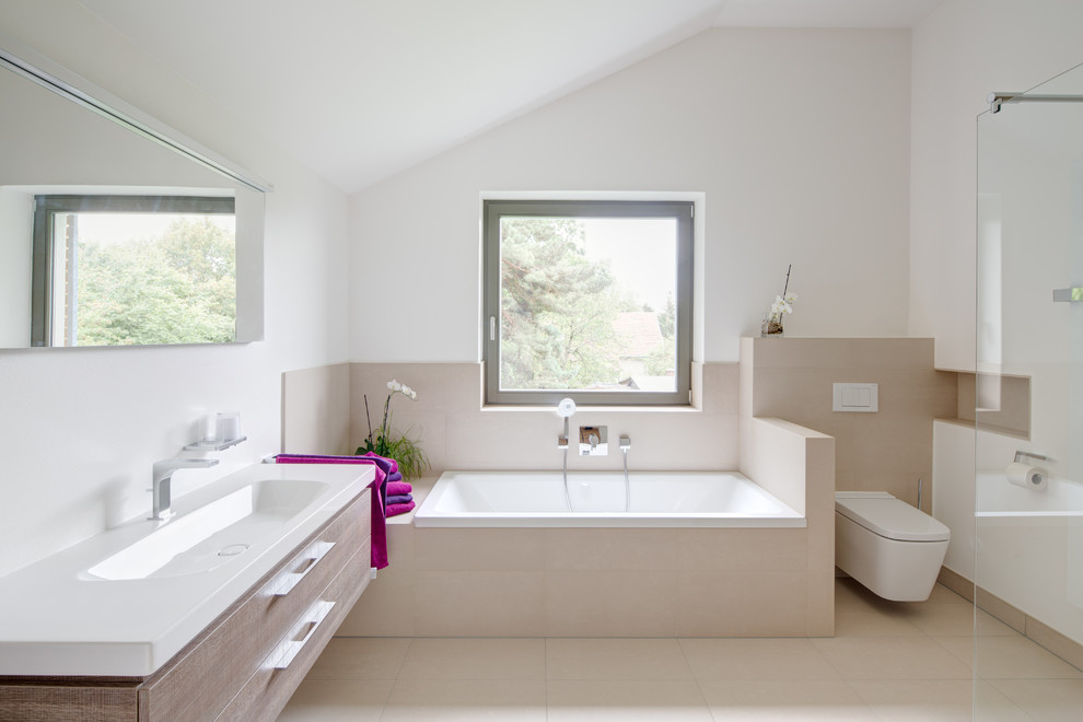На фото: большая главная ванная комната в современном стиле с плоскими фасадами, фасадами цвета дерева среднего тона, инсталляцией, бежевой плиткой, белыми стенами, ванной в нише, монолитной раковиной и окном