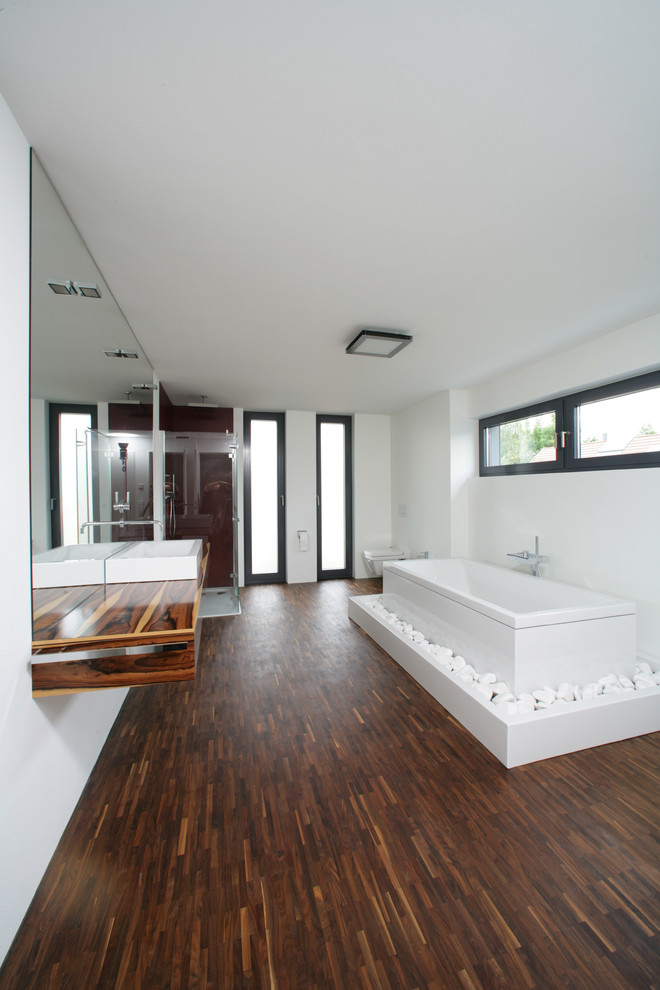 Inredning av ett modernt stort badrum med dusch, med mellanmörkt trägolv, släta luckor, skåp i mellenmörkt trä, ett fristående badkar, en öppen dusch, en vägghängd toalettstol, vita väggar, ett fristående handfat, träbänkskiva, brunt golv och dusch med gångjärnsdörr