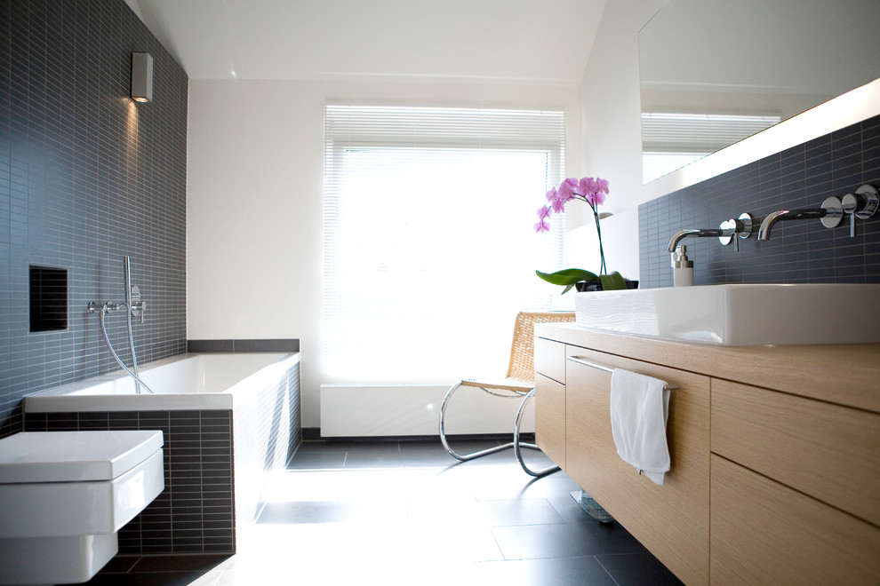 Modern inredning av ett litet brun brunt en-suite badrum, med skåp i mellenmörkt trä, ett platsbyggt badkar, en kantlös dusch, en vägghängd toalettstol, grå kakel, keramikplattor, vita väggar, klinkergolv i porslin, ett fristående handfat, träbänkskiva, grått golv och med dusch som är öppen