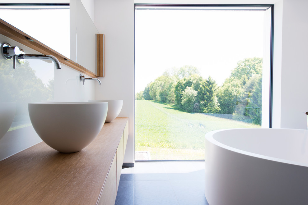 Modernes Badezimmer mit hellen Holzschränken, weißer Wandfarbe, freistehender Badewanne, grauen Fliesen und Waschtisch aus Holz in München