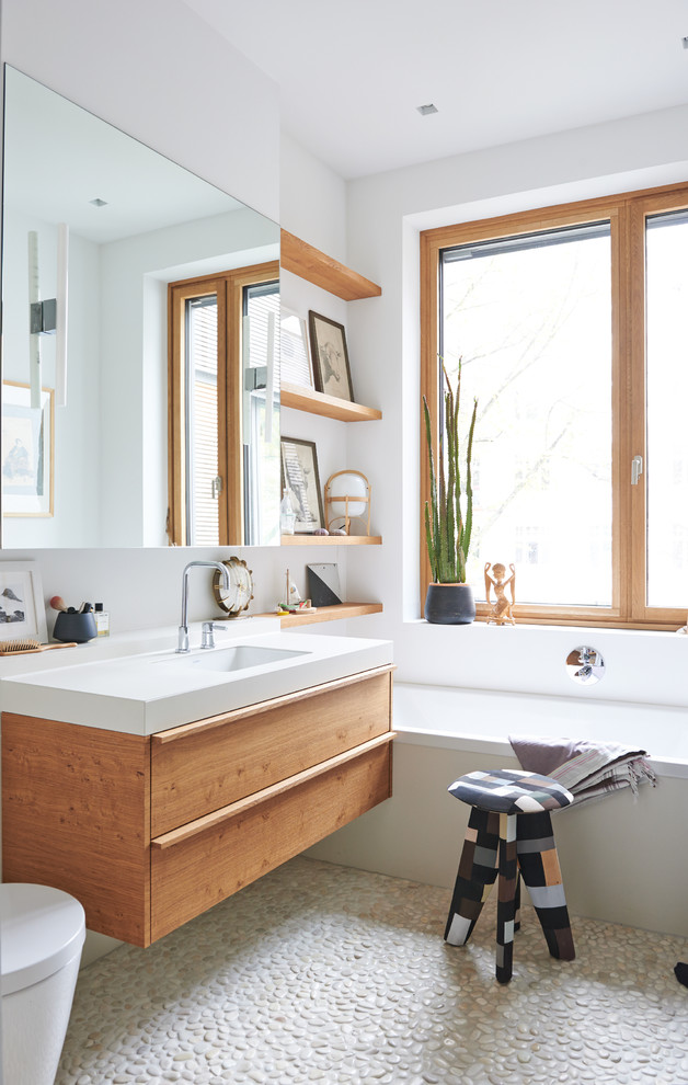 Foto på ett litet skandinaviskt badrum, med ett badkar i en alkov, vita väggar, klinkergolv i småsten, släta luckor, skåp i mellenmörkt trä och ett integrerad handfat