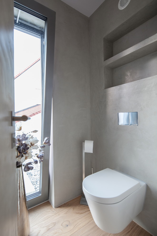 Inredning av ett modernt stort toalett, med släta luckor, vita skåp, en vägghängd toalettstol, grå kakel, cementkakel, grå väggar, ljust trägolv, ett fristående handfat, bänkskiva i betong och beiget golv