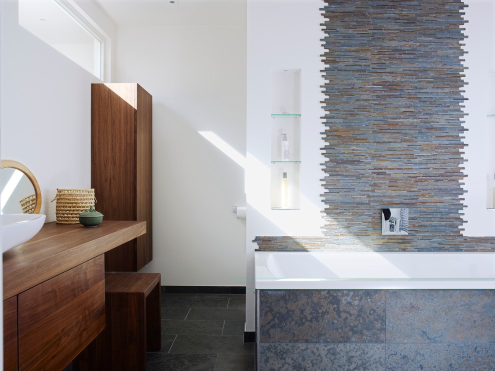Bild på ett mellanstort funkis badrum, med släta luckor, skåp i mellenmörkt trä, ett platsbyggt badkar, flerfärgad kakel, stickkakel, vita väggar, ett fristående handfat och träbänkskiva