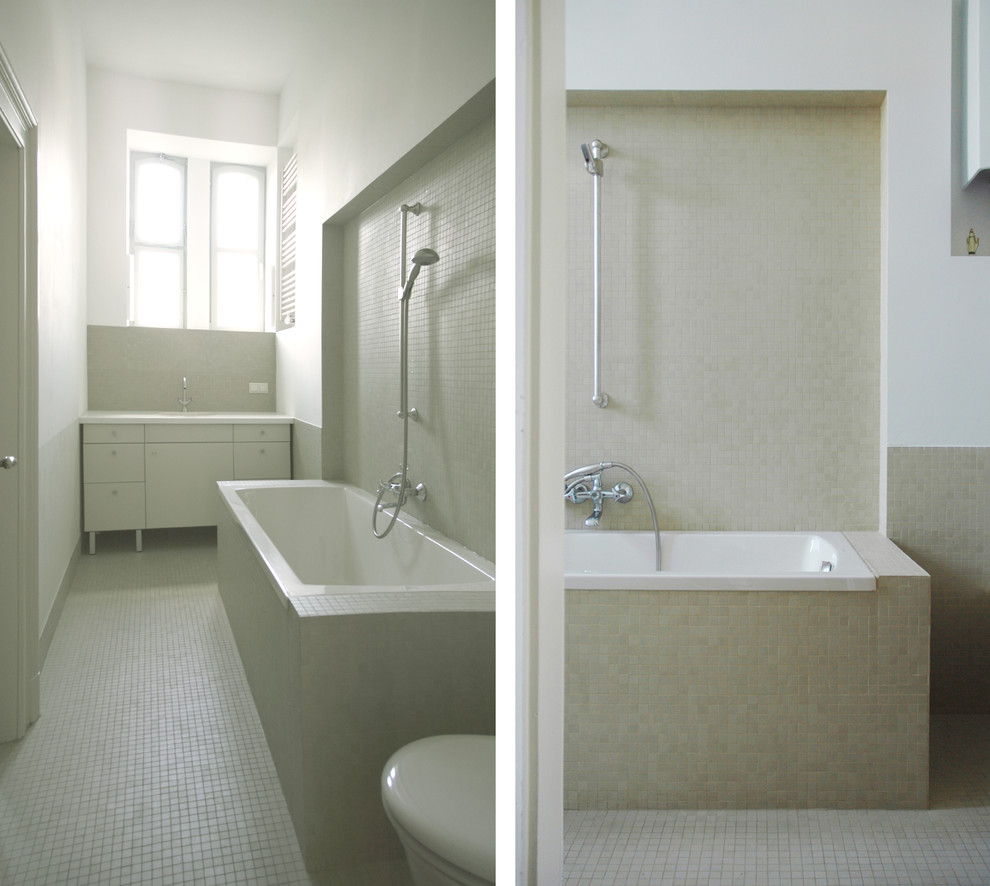 Bild på ett lantligt badrum, med ett nedsänkt handfat, släta luckor, vita skåp, laminatbänkskiva, ett platsbyggt badkar, en toalettstol med separat cisternkåpa, vit kakel, porslinskakel, vita väggar och mosaikgolv