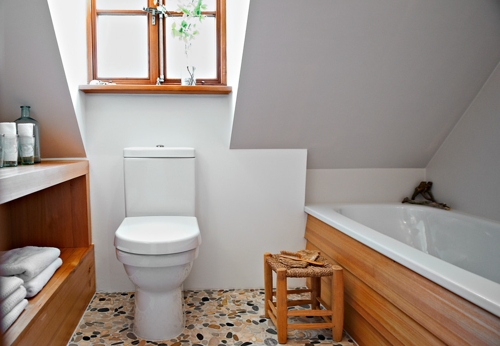 Cette photo montre une petite salle de bain nature en bois brun avec un placard sans porte, une baignoire posée, un mur blanc, un sol en galet, une plaque de galets et WC à poser.