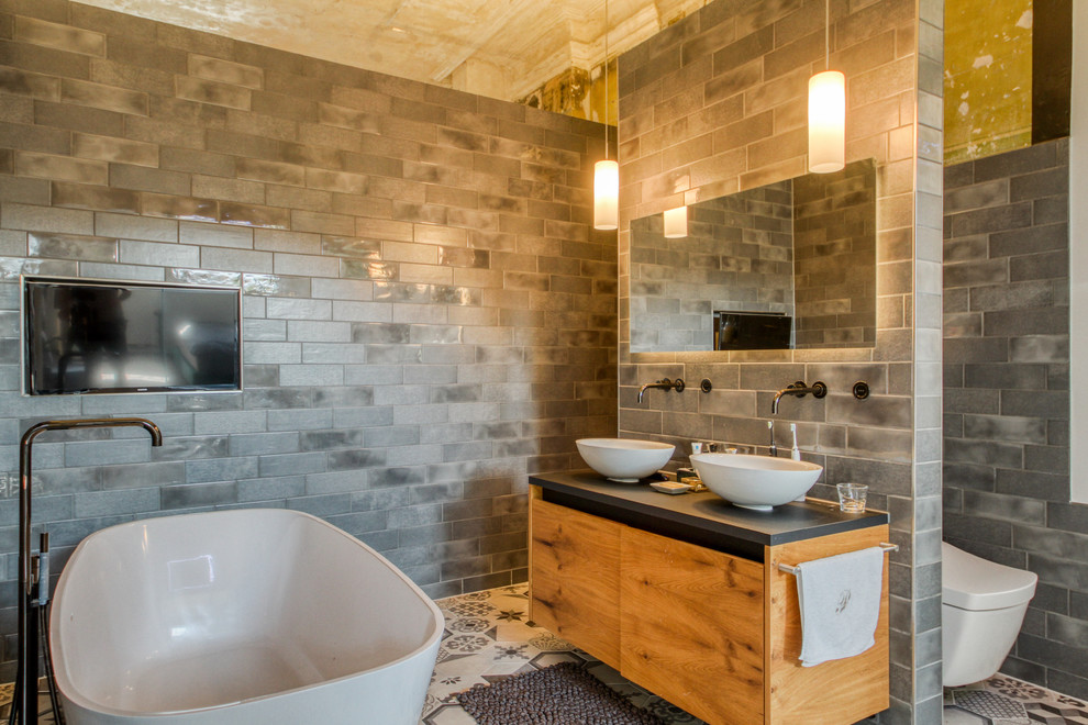 This is an example of a contemporary ensuite bathroom in Stuttgart with flat-panel cabinets, medium wood cabinets, a freestanding bath, a wall mounted toilet, grey tiles, a vessel sink, grey floors and black worktops.