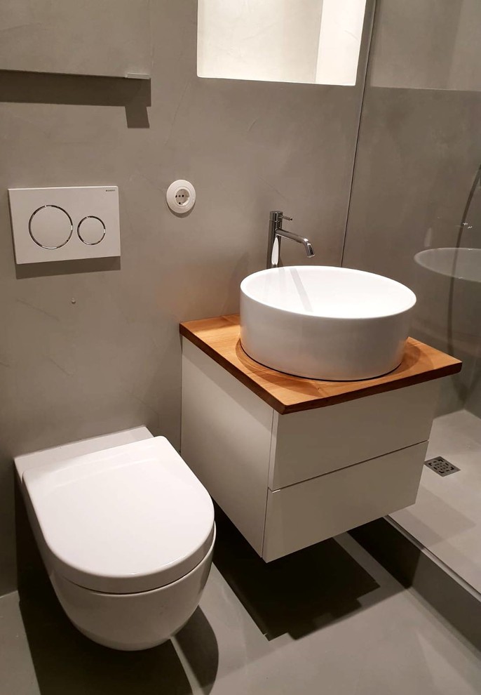 Modern inredning av ett litet brun brunt badrum med dusch, med luckor med profilerade fronter, vita skåp, en öppen dusch, en vägghängd toalettstol, grå väggar, betonggolv, ett fristående handfat, träbänkskiva, grått golv och med dusch som är öppen