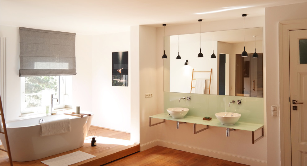 Modernes Badezimmer mit Aufsatzwaschbecken, Unterbauwaschbecken, freistehender Badewanne, weißer Wandfarbe, braunem Holzboden und Glaswaschbecken/Glaswaschtisch in Düsseldorf