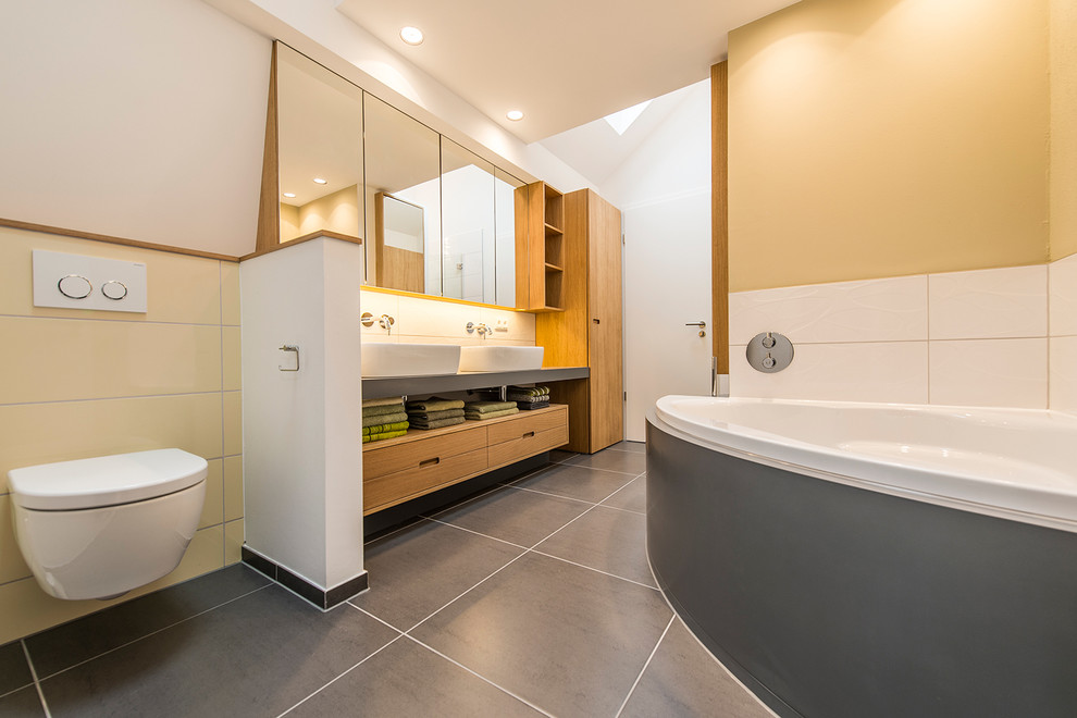 Inredning av ett modernt mellanstort badrum, med ett fristående handfat, skåp i mellenmörkt trä, bänkskiva i akrylsten, ett hörnbadkar, en kantlös dusch, en vägghängd toalettstol, grå kakel, gul kakel, vit kakel, keramikplattor, vinylgolv och gula väggar