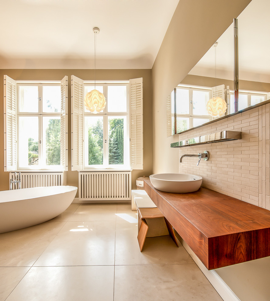 Cette image montre une grande salle de bain design avec une vasque, un plan de toilette en bois, une baignoire indépendante, un carrelage beige, un mur beige, des carreaux en allumettes et un plan de toilette marron.