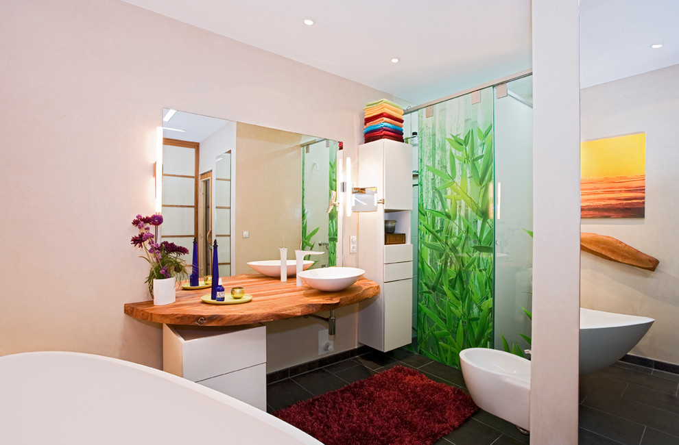 Foto på ett mellanstort funkis en-suite badrum, med släta luckor, vita skåp, ett fristående badkar, en kantlös dusch, en bidé, svart kakel, stenkakel, beige väggar, ett fristående handfat och träbänkskiva