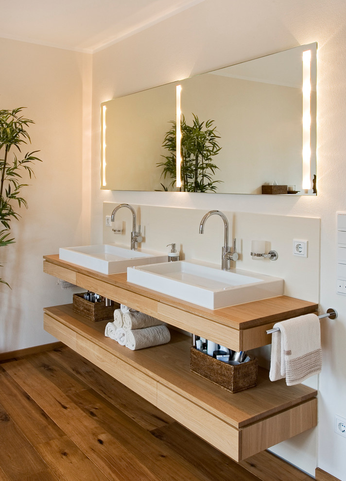 Foto på ett mellanstort funkis brun en-suite badrum, med släta luckor, ett fristående badkar, stenkakel, beige väggar, mellanmörkt trägolv, ett fristående handfat, träbänkskiva, skåp i ljust trä och beige kakel
