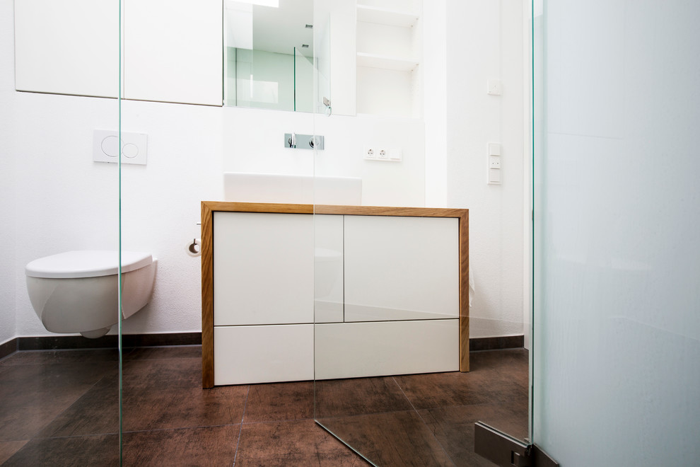 Design ideas for a medium sized contemporary ensuite bathroom in Munich with a wall mounted toilet, flat-panel cabinets, white cabinets, a built-in bath, a built-in shower, white tiles, porcelain tiles, white walls, cement flooring, a vessel sink, wooden worktops, brown floors and an open shower.