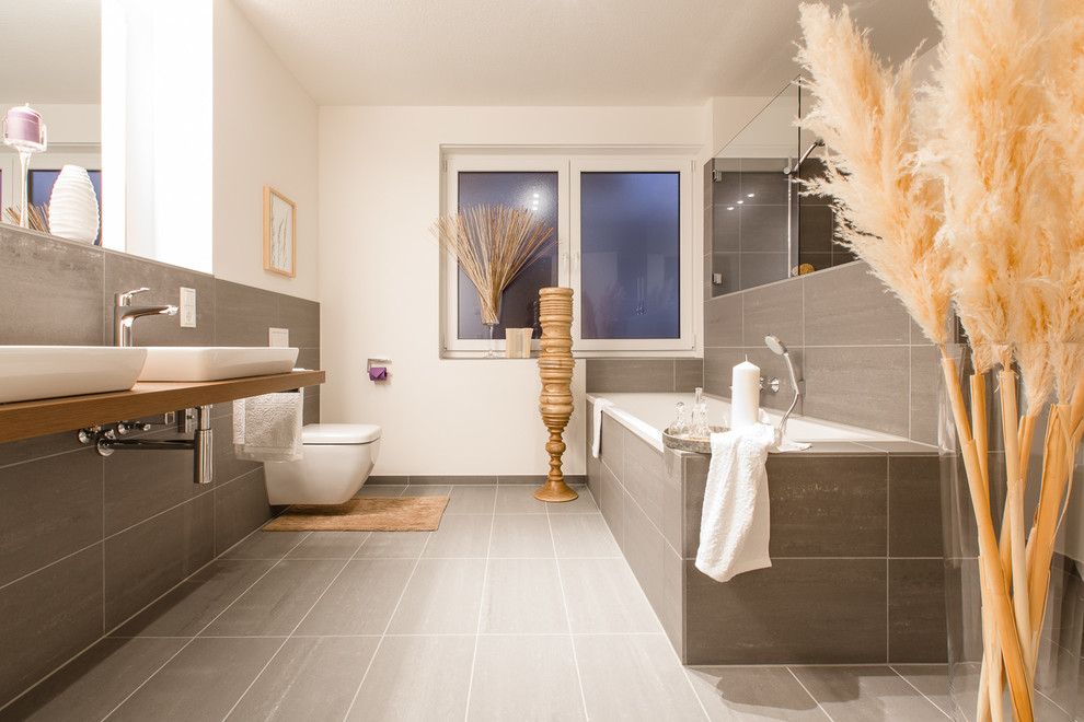 Inredning av ett modernt stort badrum, med ett platsbyggt badkar, en vägghängd toalettstol, grå kakel, stenkakel, vita väggar, ett fristående handfat, träbänkskiva och en dusch i en alkov